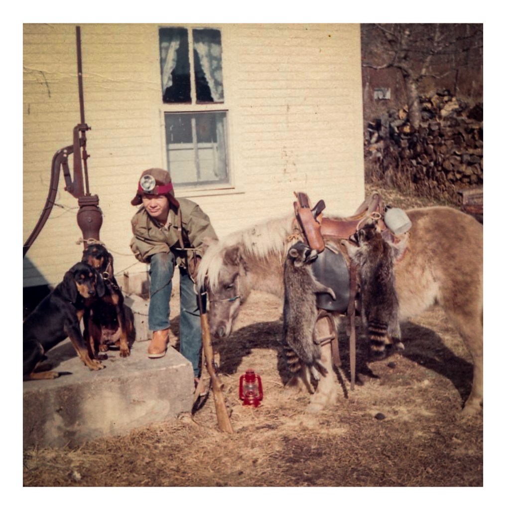 Young man leans on concrete step next to two black and tan dogs and a pale sheltland pony.